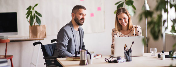 business_man_in_wheelchair.wearing_suit_sits_at_a_large_desk_in_meeting_with_blonde_female_colleague_who_has_a_laptop