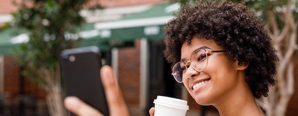 Social media user smiles at camera phone as she holds it one hand with a coffee cup in the other