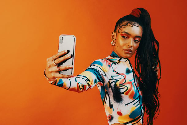 Mulher posa para selfie. Fundo laranja contrasta com cores da roupa e maquiagem. Instagram aesthetic. 