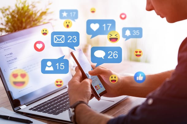imagem com pessoa checando redes sociais em seu celular na frente de um computador mostra ícones que representam reações, emojis, comentários e likes em diferentes plataformas de redes sociais 