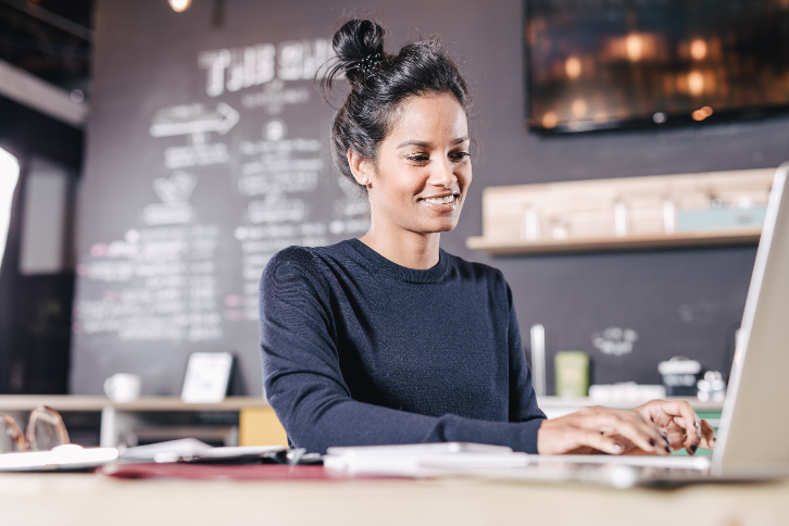 mulher sentada em uma cafeteria digita conteúdo em um laptop. Ela está sorrindo. 